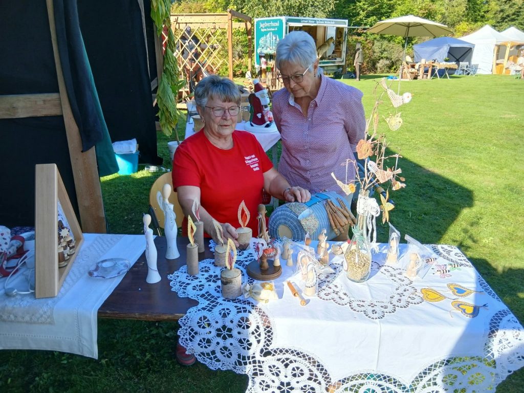 Zwei Vereinsmitglieder an der Klöppelrolle, dahinter der Tisch mit Schnitzarbeiten