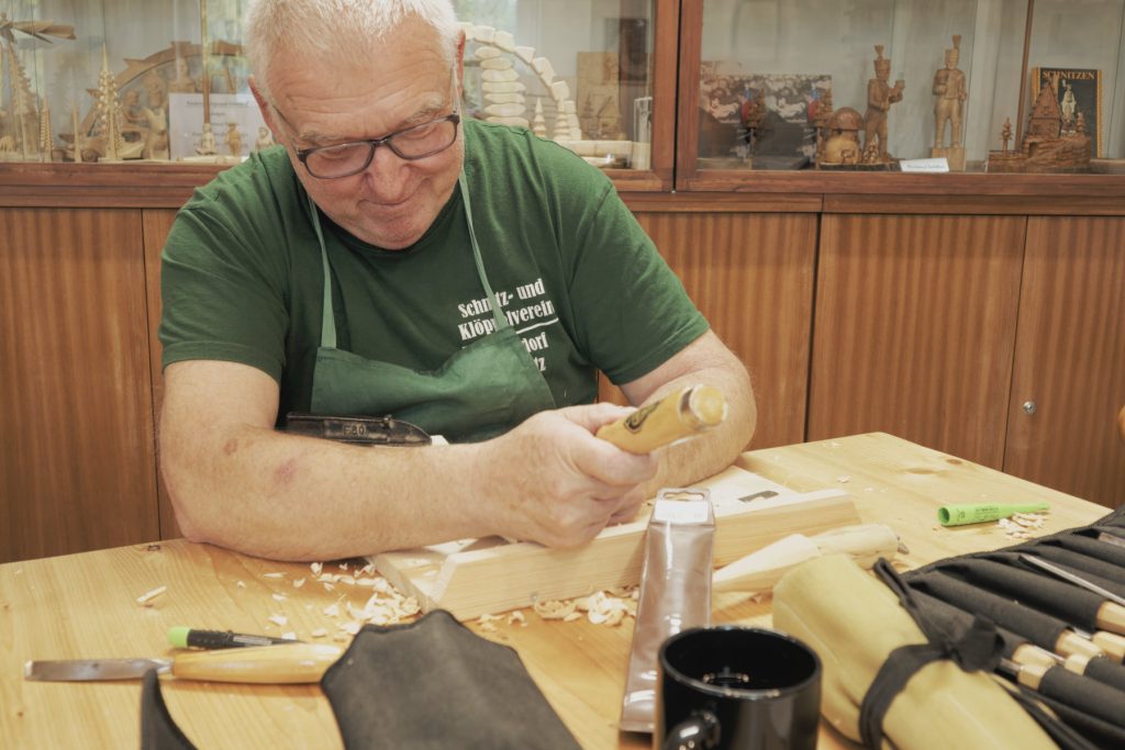 Vereinsmitglied schnitzt mit Klüpfel und Eisen an einem Holzstück, Späne auf dem Tisch