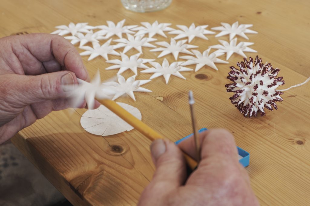Hände formen weiße Papiersterne zu einer Kugel, daneben eine fertige Kugel mit roter Kante